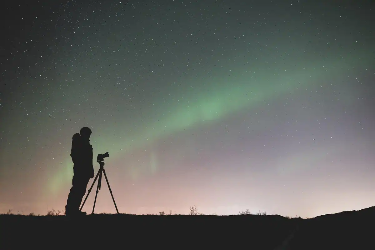 Siluetă de fotograf cu trepied sub o aurora verzuie pe cerul nopții