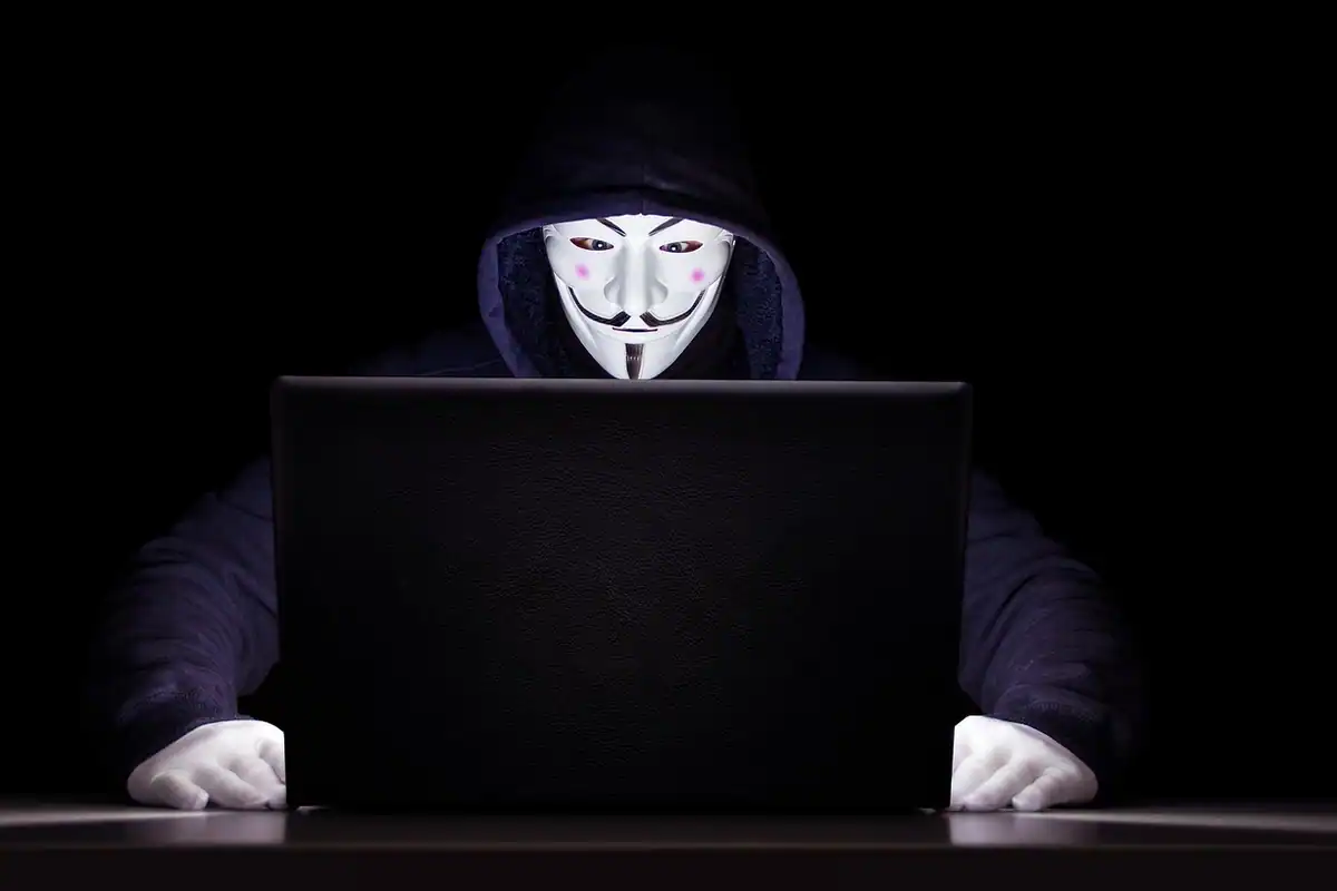 Persoană cu glugă și mască albă în fața unui laptop, într-o cameră întunecată