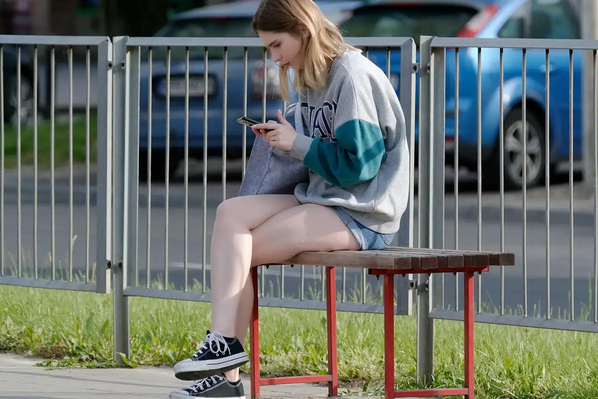Tânără așezată pe o bancă, care se uită la telefon pe stradă