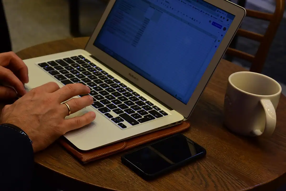 Mâini pe tastatura unui laptop, cu o cană și un telefon alături